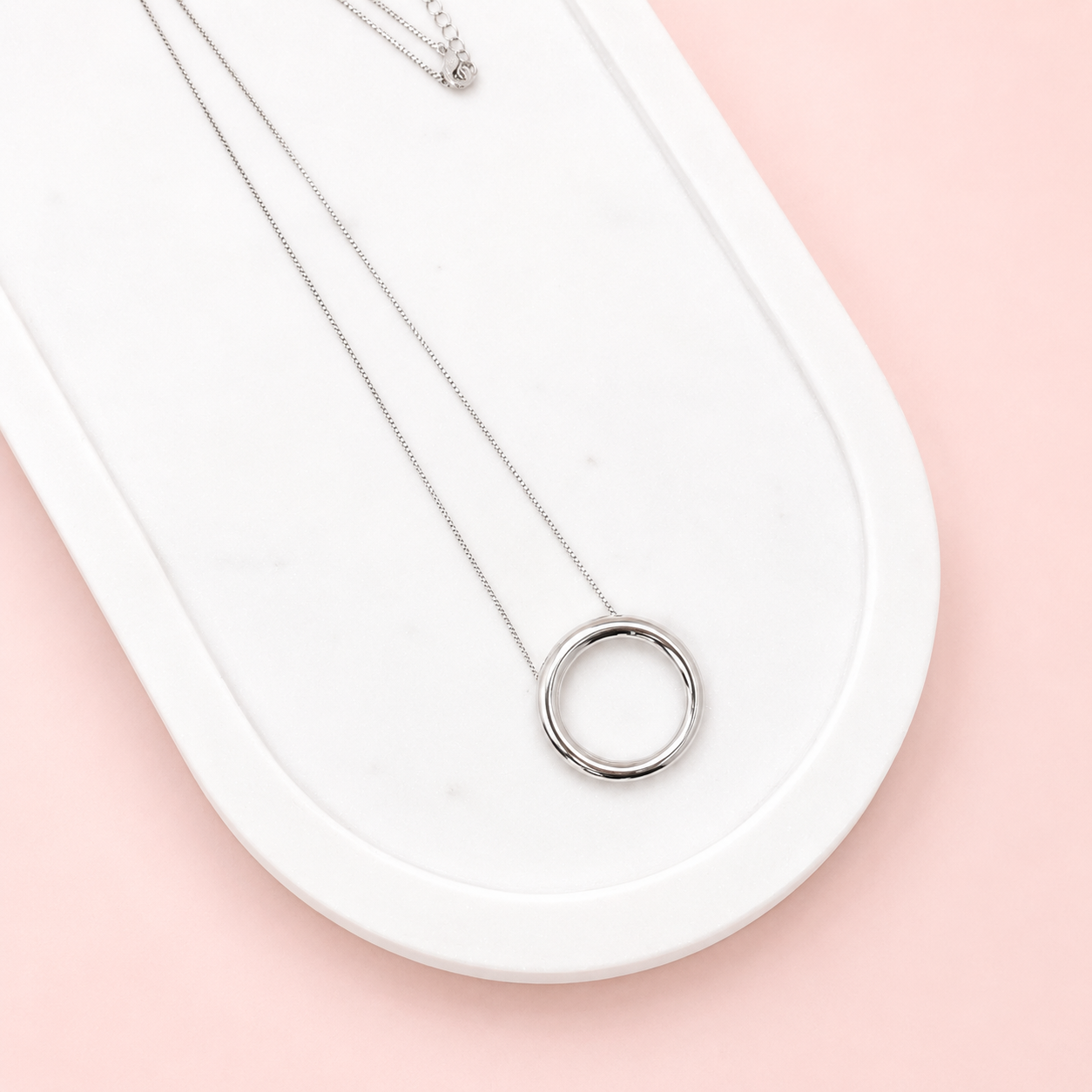 Silver necklace with a circular pendant on a white surface with a pink background